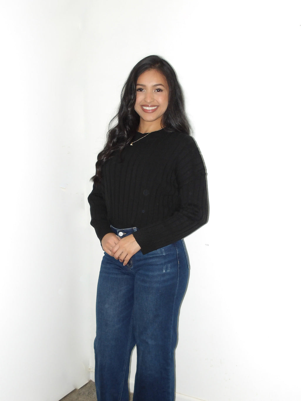 Woman wearing a black sweater and blue jeans standing against a white wall.